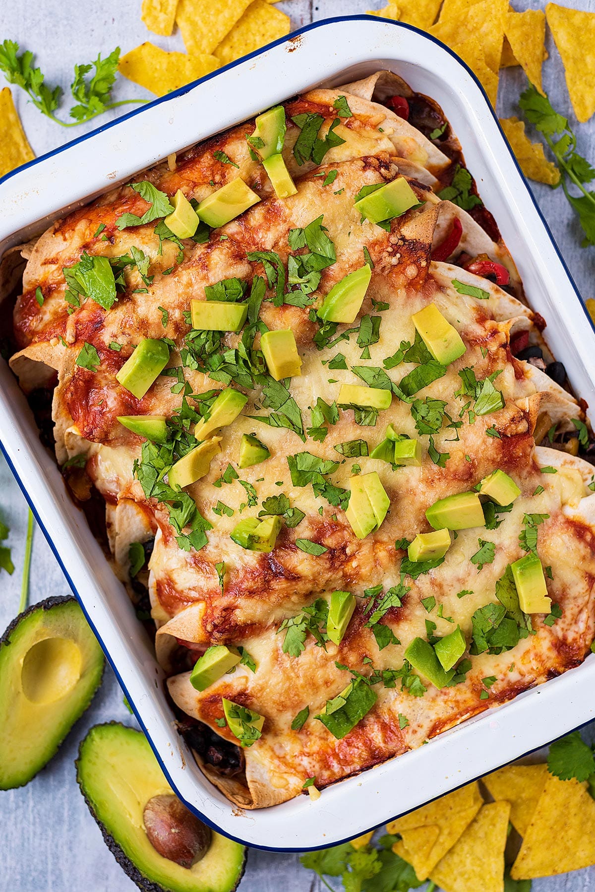 A baking dish full of enchiladas next to tortilla chips and avocado halves.