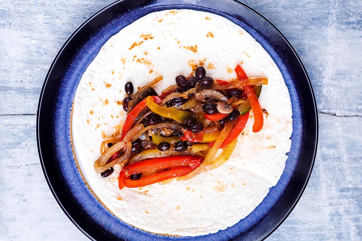 A flour tortilla with cooked beans and sliced vegetables along the middle of it.