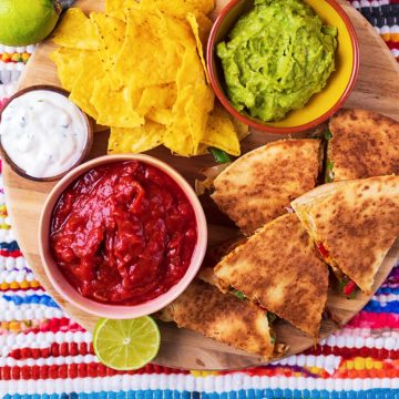 Slices of chicken quesadilla on a serving board with tortilla chips, salsa and guacamole