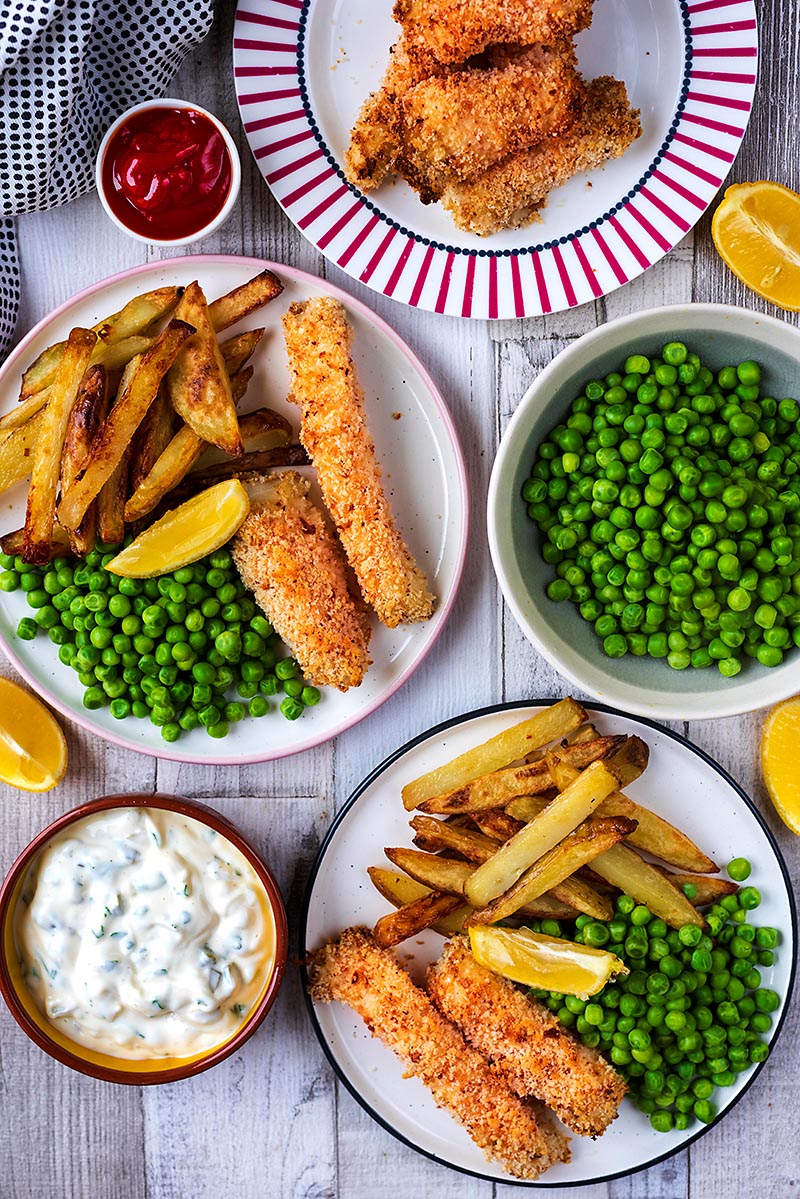 Two plates with fish sticks, fries and peas next to a bowl of tartar sauce.