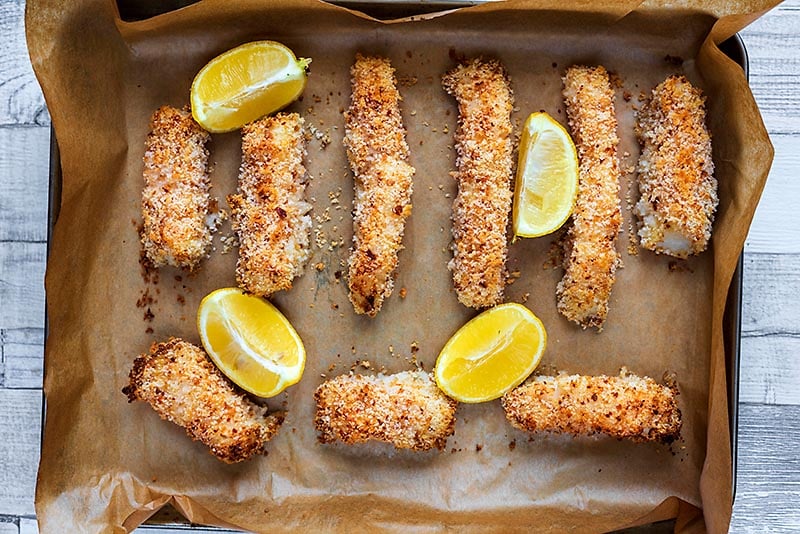 Nine cooked fish sticks on a baking tray with four lemon wedges.