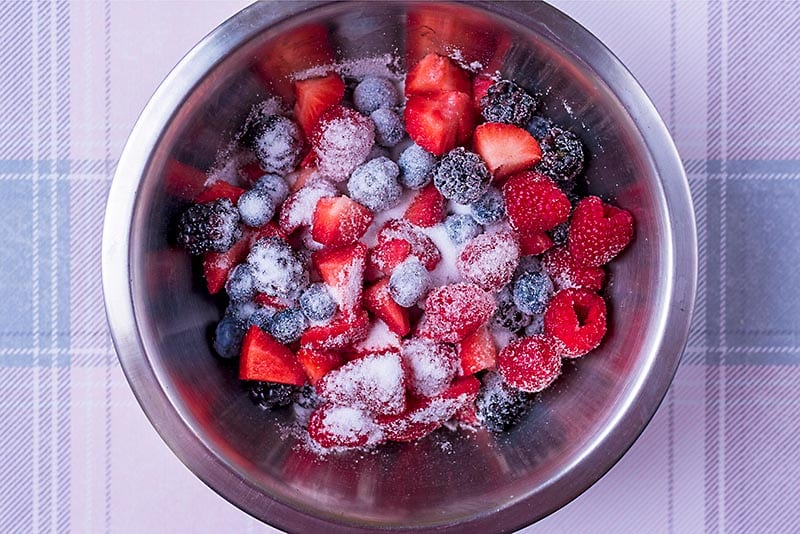 A large stainless steel bowl containing mixed berries and sugar