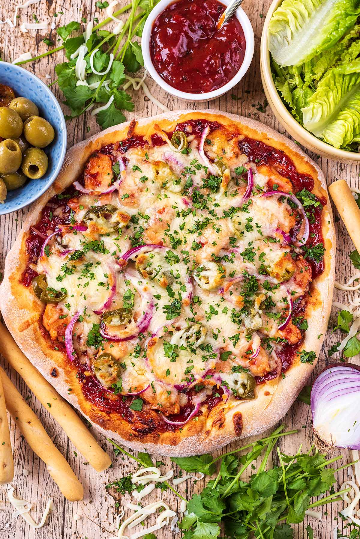 A thick crust pizza surrounded by salad, olives, bread sticks, herbs and dip.