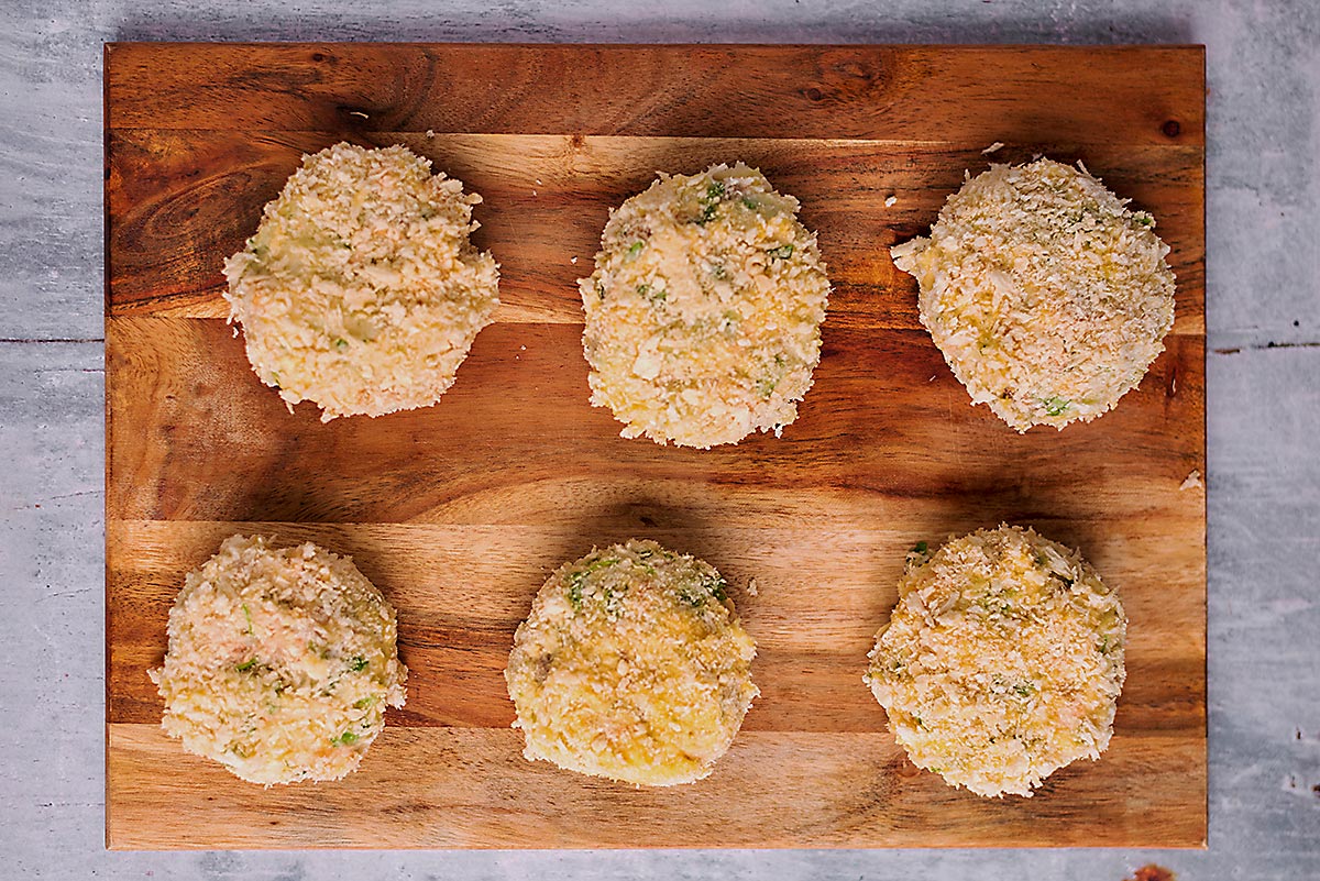 Six uncooked breaded fishcakes on a wooden board.