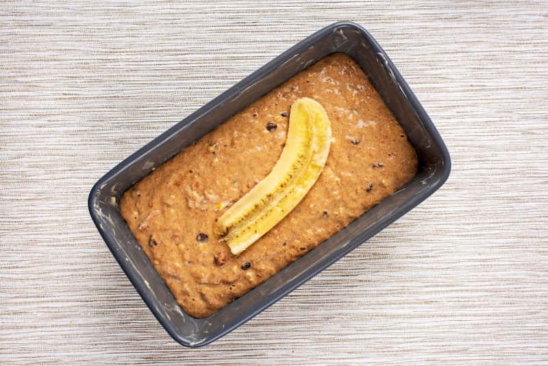A loaf tin containing a brown cake batter topped with sliced banana.