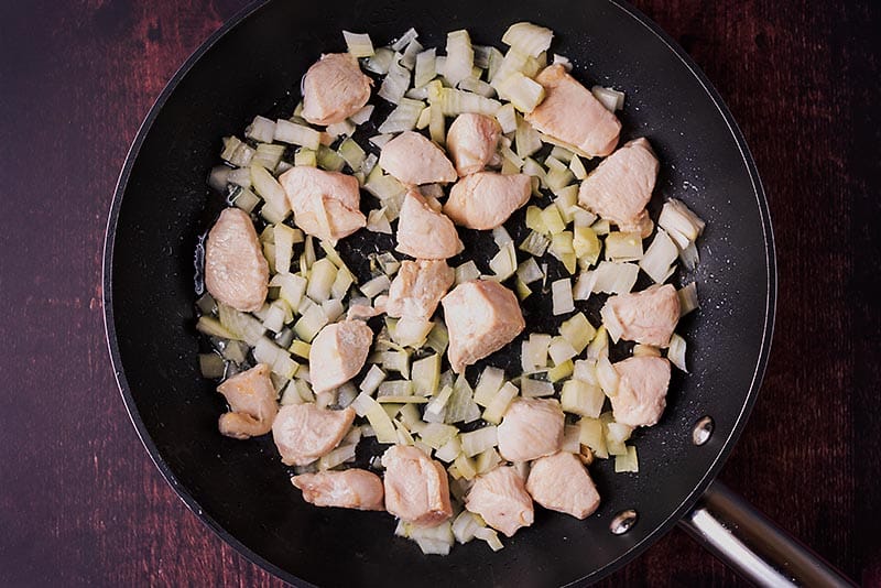 Chunks of chicken breast and chopped onions cooking in a frying pan.
