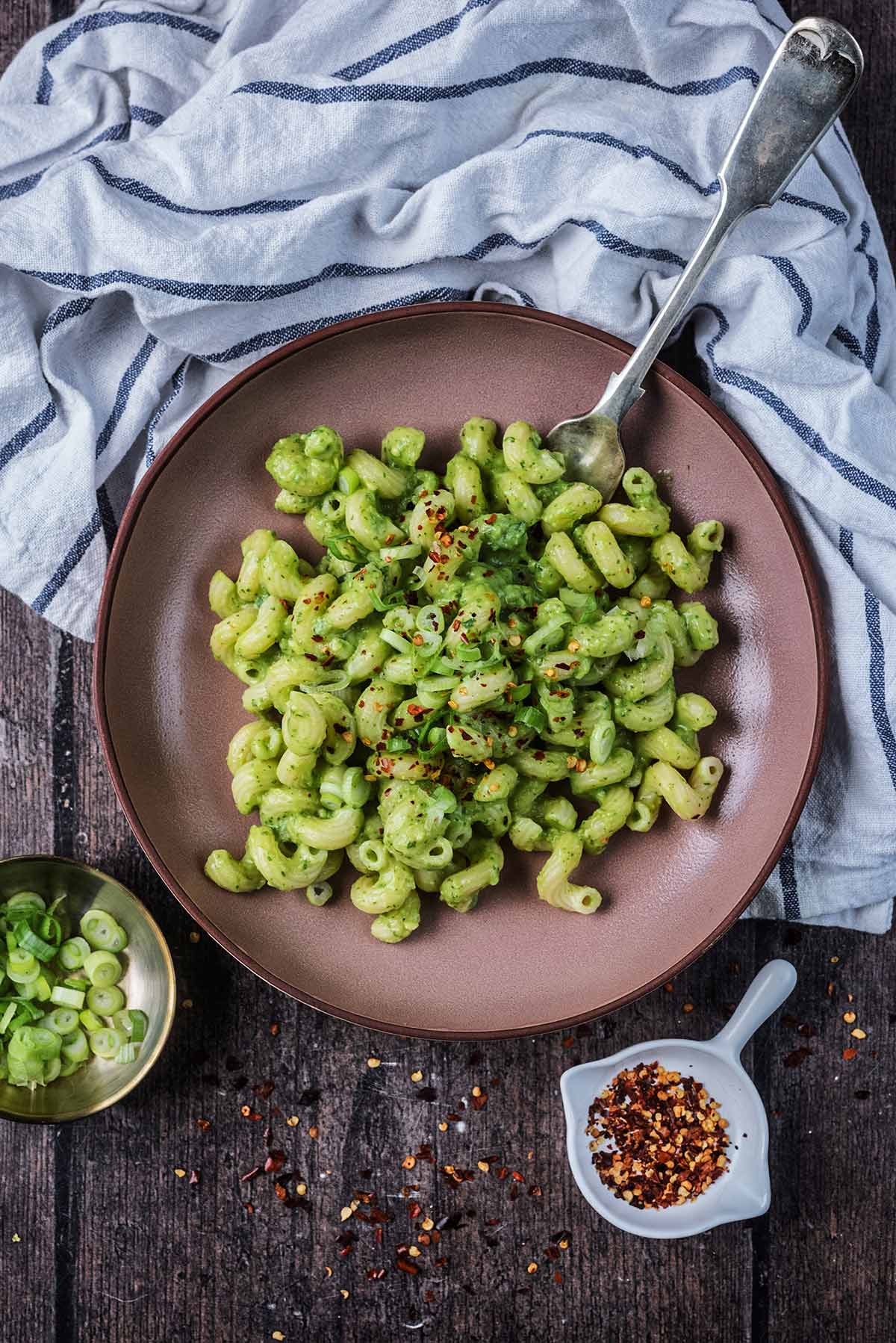 A large bowl of cooked pasta in agreen sauce next to chopped spring onions and red chilli flakes.