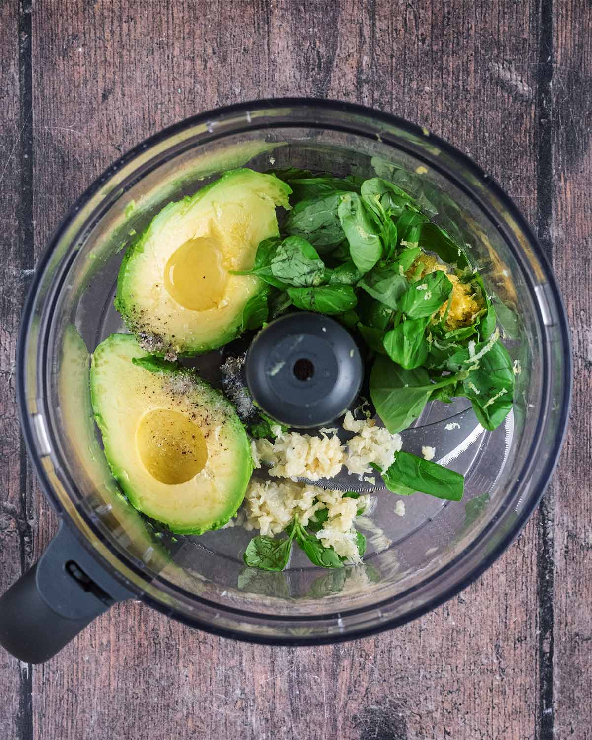 A food processor containing avocado, basil leaves, garlic cloves, lemon zest and seasoning.