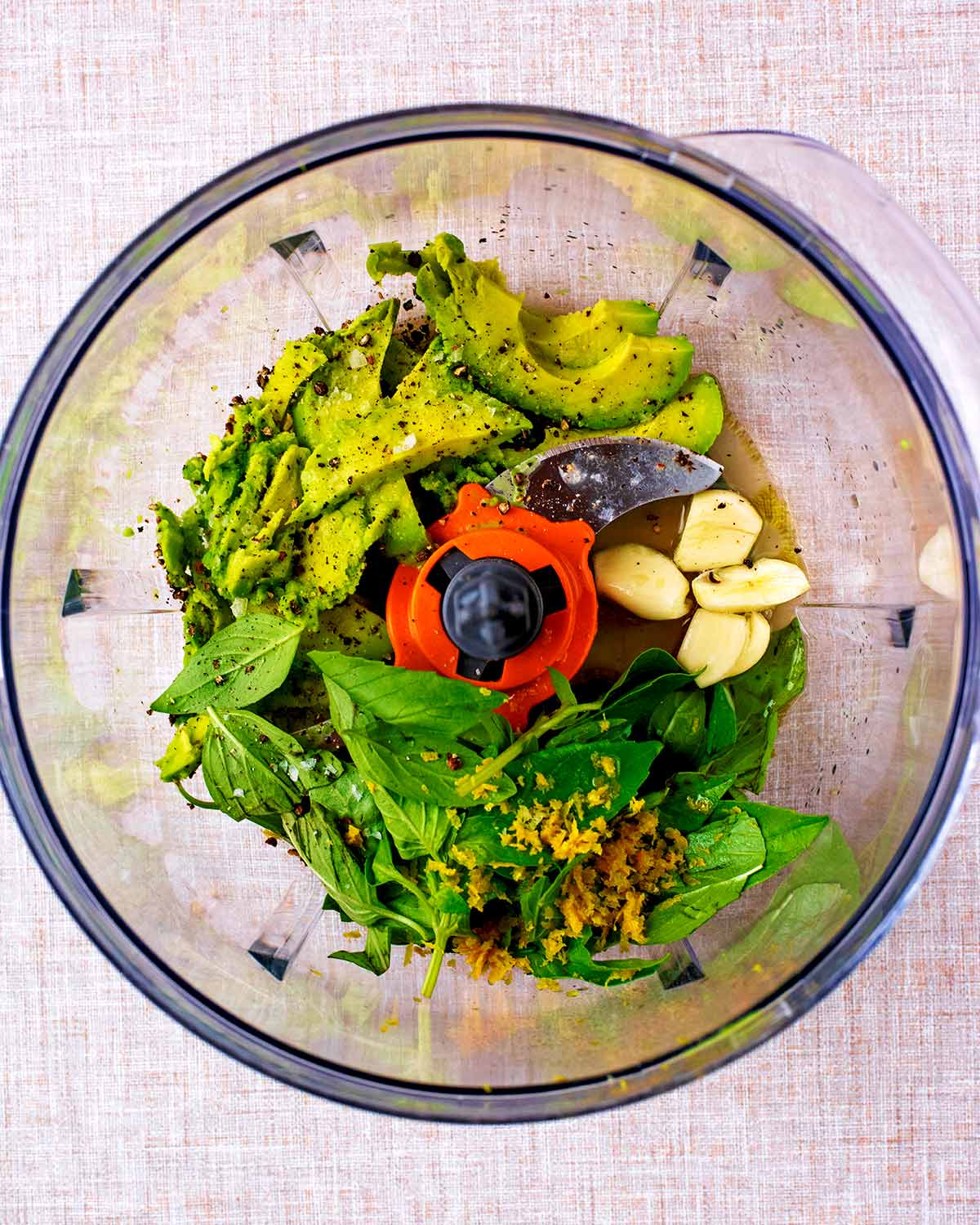 A food processor containing avocado, basil leaves, garlic cloves, lemon zest and seasoning.