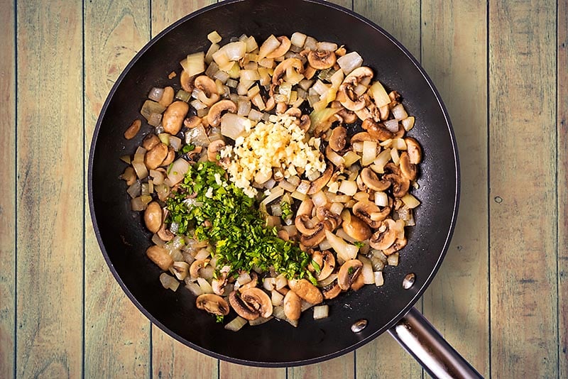 A frying pan containing cooked onions and mushrooms with chopped herbs and minced garlic.