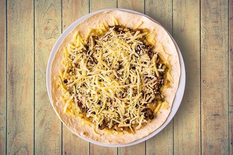 A flour tortilla covered in cooked mushroom and onions and then covered in grated cheese.
