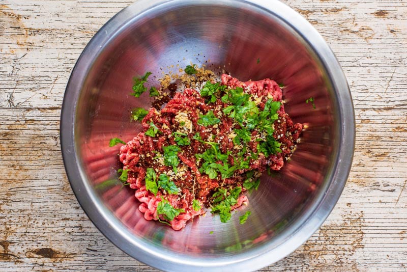 A mixing bowl containing raw ground beef, herbs and spices.