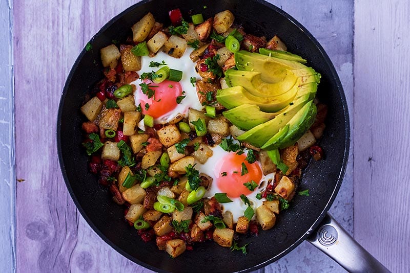 Breakfast Hash in a frying pan with two cooked eggs and sliced avocado.