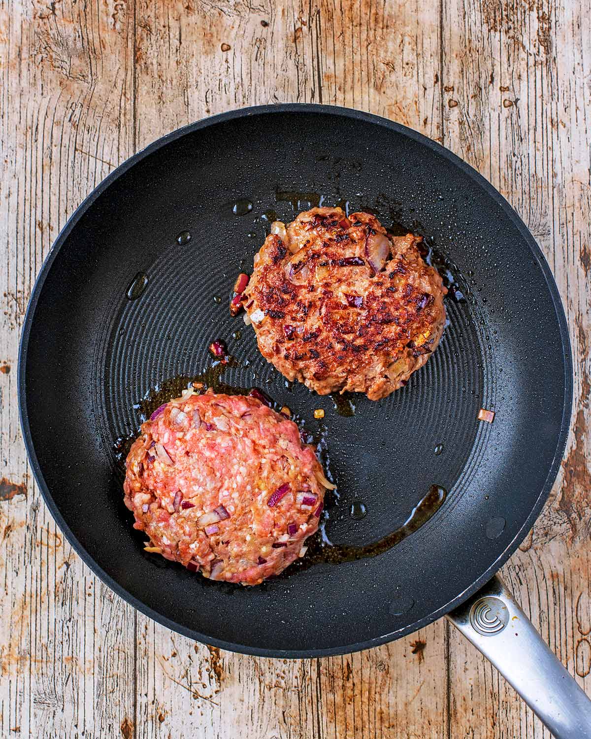 A frying pan with two burgers cooking in it. One has been flipped over to show the cooked side.