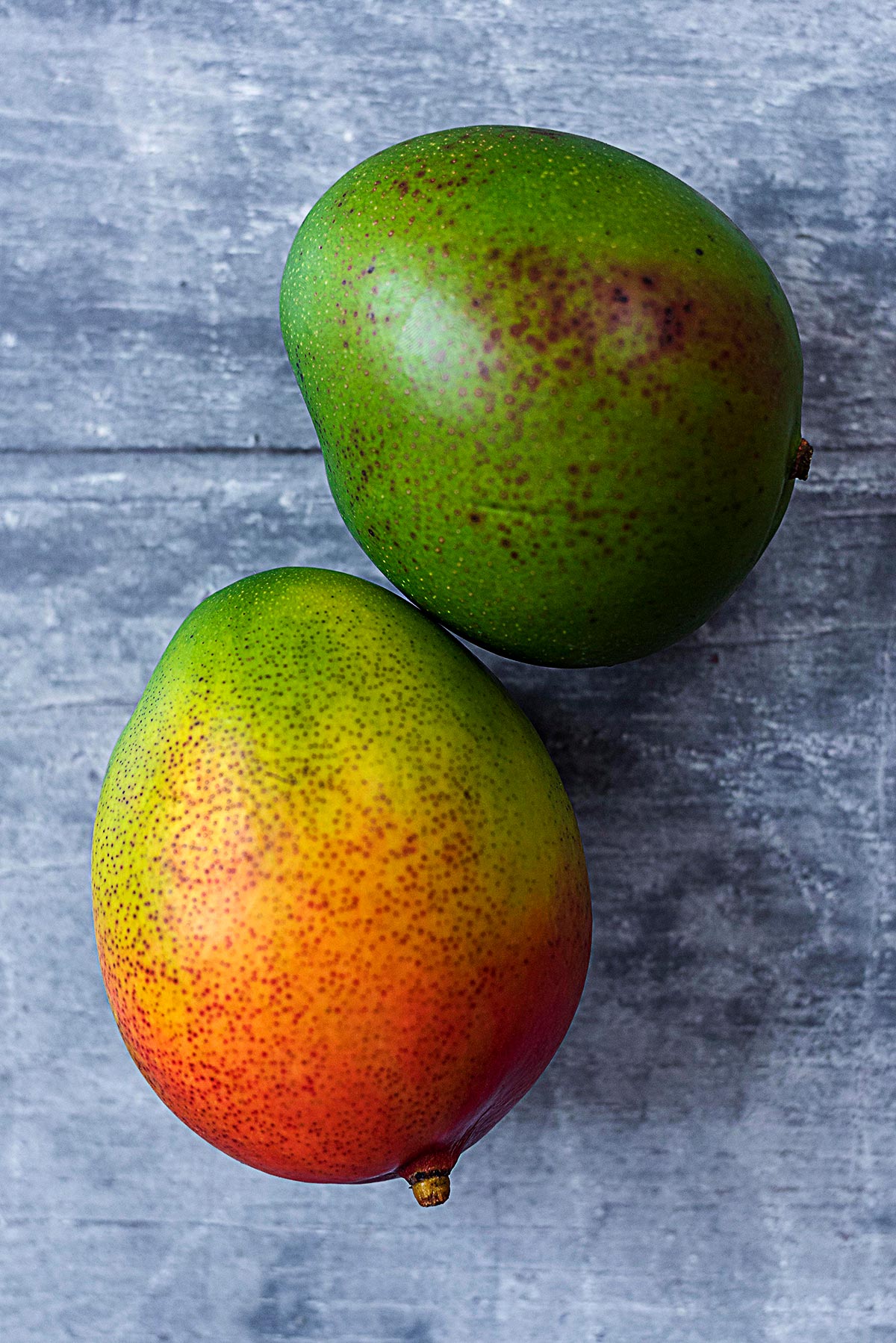 Two mangoes on a wooden surface. One is all green, one is green and red.
