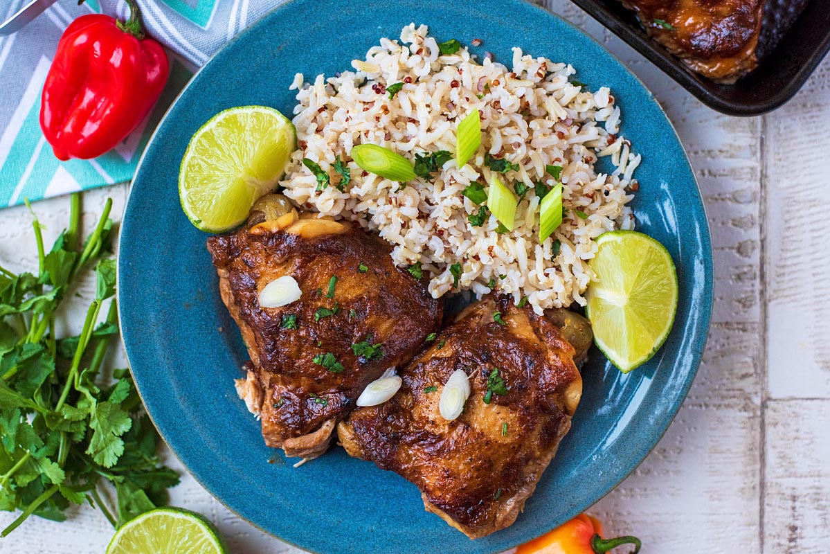 Two jerk chicken thighs on a blue plate with rice and lime wedges.