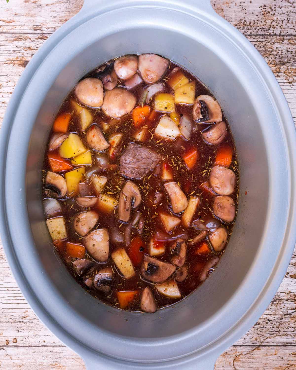 A slow cooker bowl containing chunks of beef, chopped vegetables and gravy.