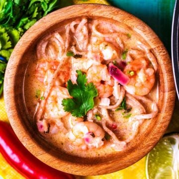Prawn Laksa in a wooden bowl next to a bunch of cilantro, a red chili and a lime wedge