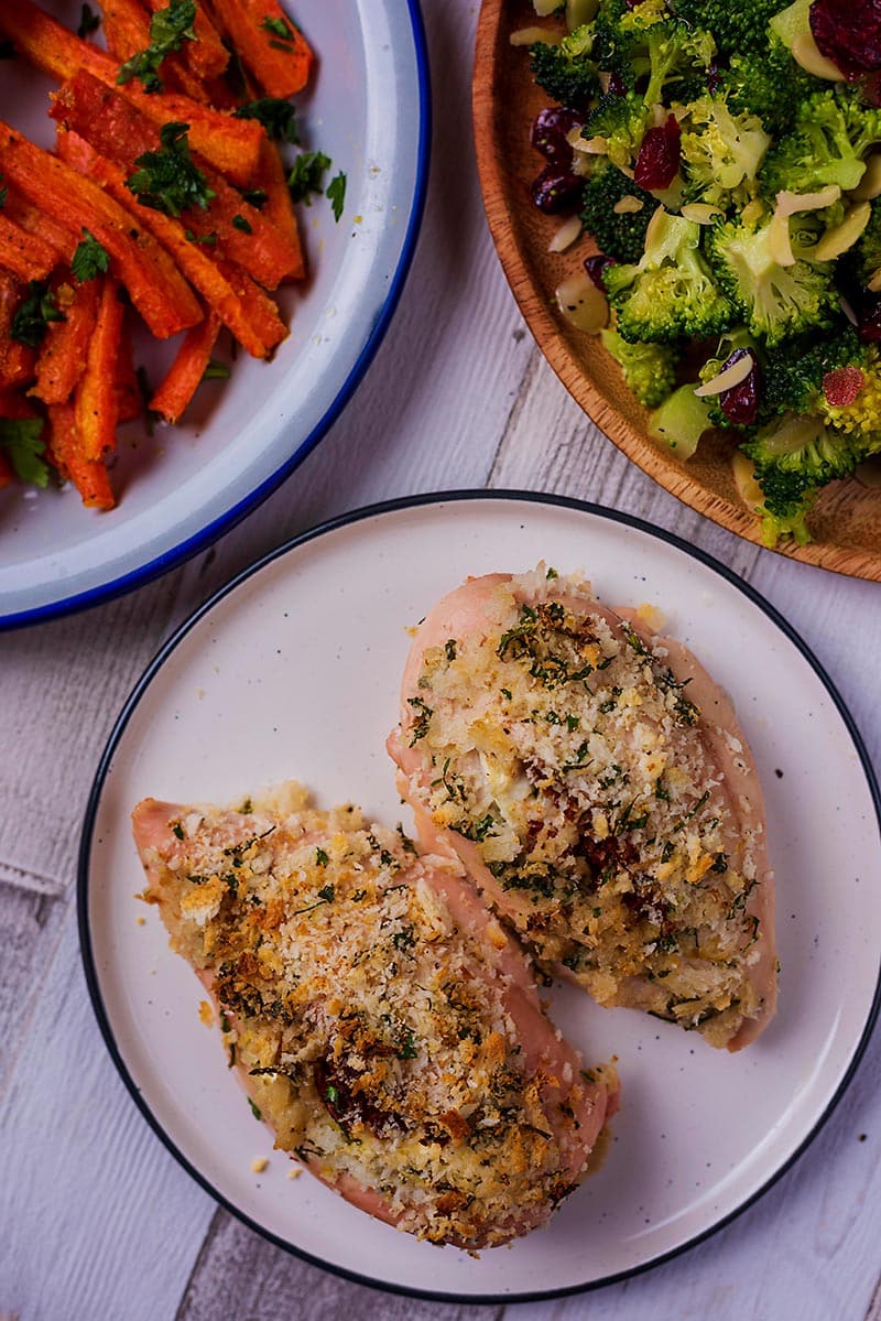 Two stuffed chicken breasts on a plate next to plates of carrots and broccoli.