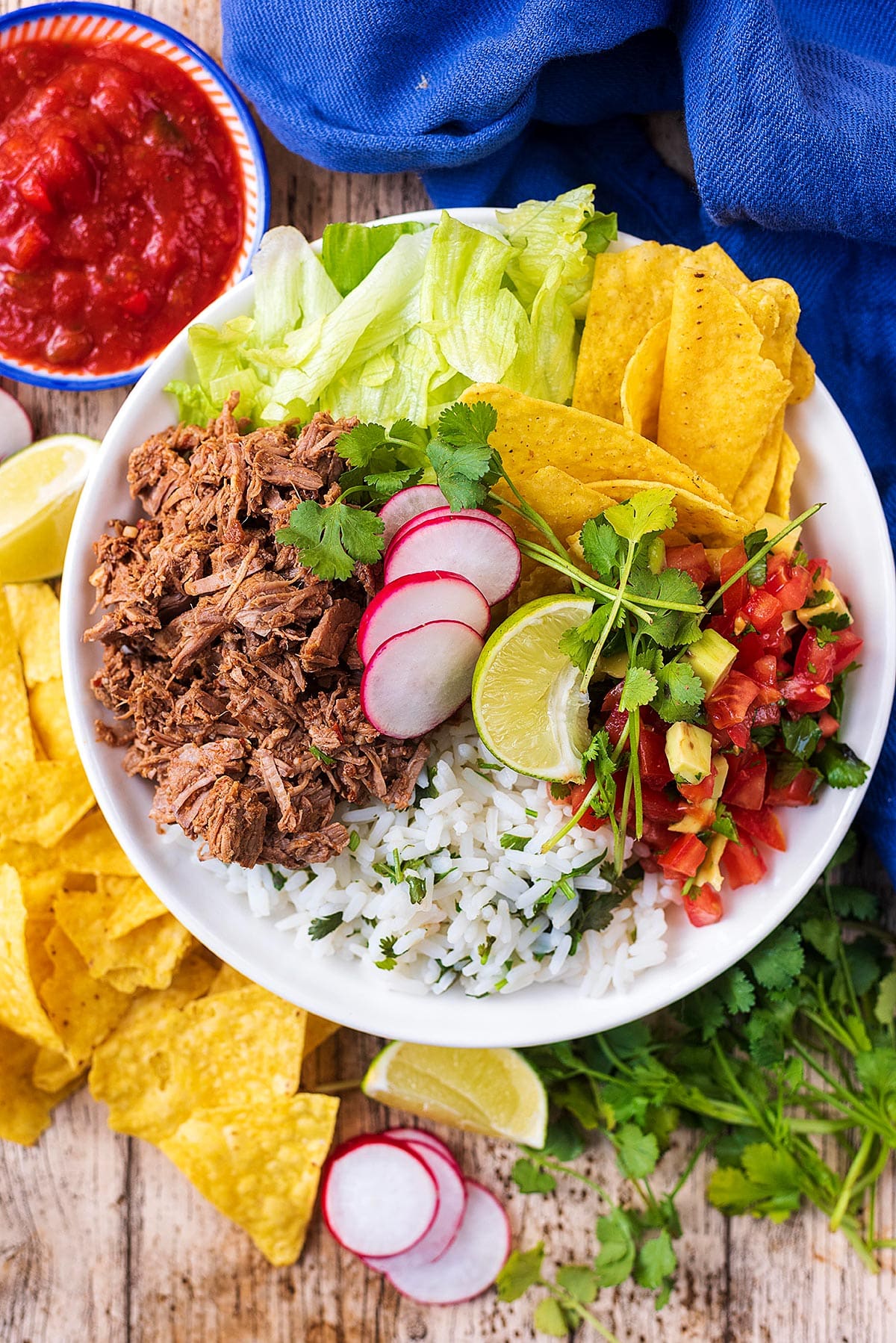 A burrito bowl consisting of shredded beef, lettuce, salsa, rice, tortilla chips and sliced radish.