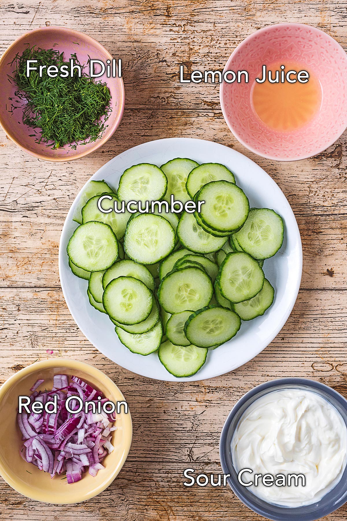 A bowl of sliced cucumber surrounded by bowls of dill, lemon juice, chopped onion and sour cream.