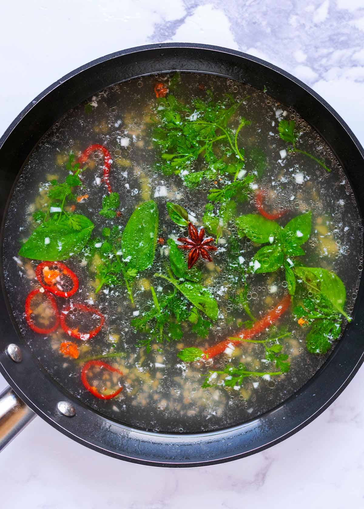 A frying pan containing a broth with herbs, chillies and spices.