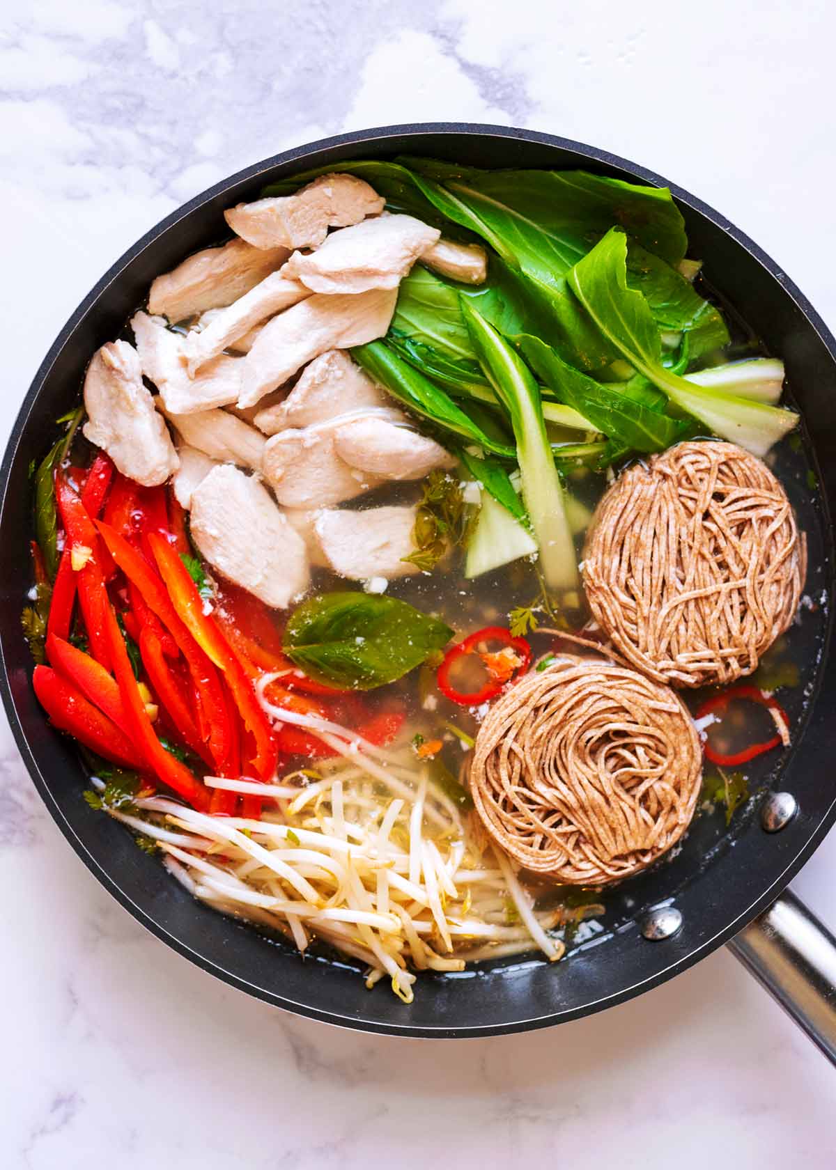 A pan containing broth, chicken, sliced peppers, beansprouts, bok choi and noodles.