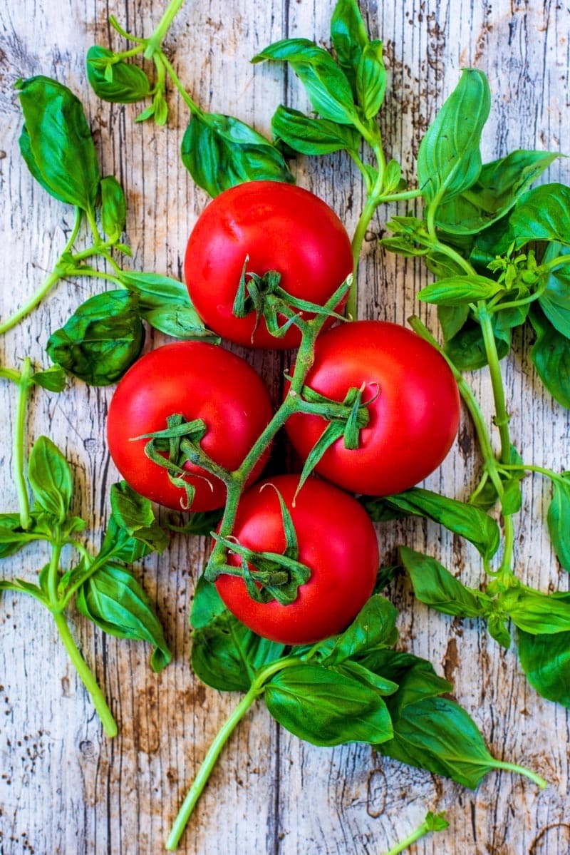 Four ripe tomatoes on the vine with basil leaves all on a wooden surface.