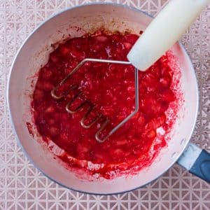 A saucepan with mashed strawberries and a potato masher