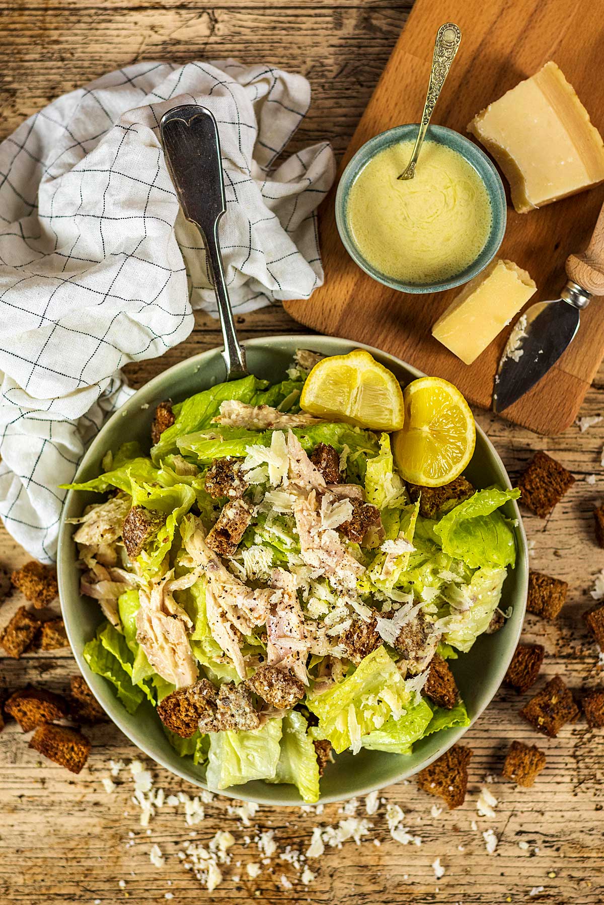 A large bowl of caesar salad next to a chopping board with cheese, a knife and a bowl of caesar dressing.