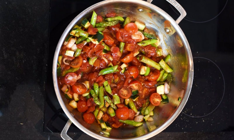 Chopped vegetables and chopped tomatoes cooking in a large silver pan.