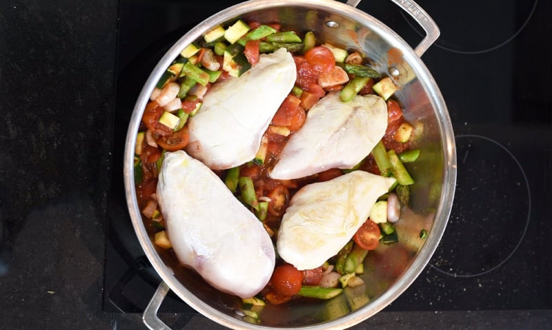 Four part cooked chicken breasts on top of cooked vegetables in a large silver pan.
