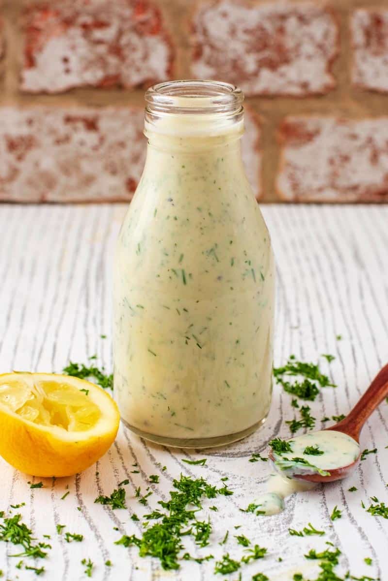 A glass bottel of healthy ranch dressing next to a lemon half and a small wooden spoon.