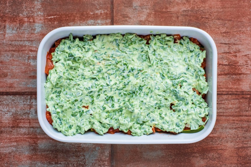 A baking dish containing a tomato meat sauce, strips of cooked zucchini and a yoghurt spinach sauce.