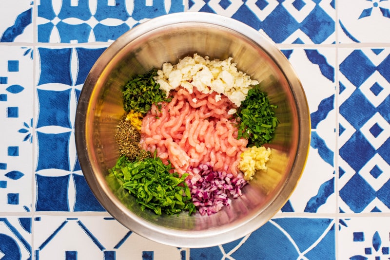 A metal mixing bowl with ground turkey, minced garlic, feta, herbs and spices.