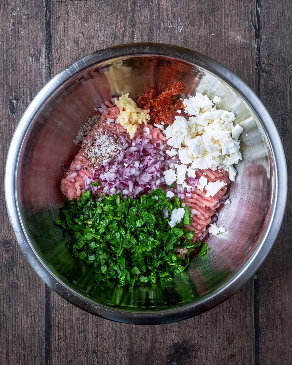 A metal mixing bowl with ground turkey, minced garlic, feta, herbs and spices.