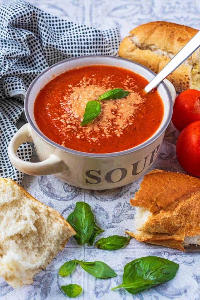 A bowl of tomato soup in a cream soup bowl. Bread and basil leaves are scattered around