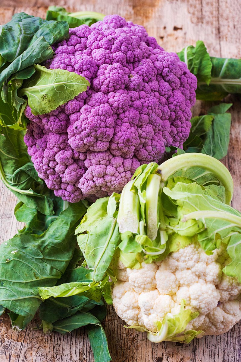 A whole white cauliflower and a whole purple cauliflower on a wooden board.