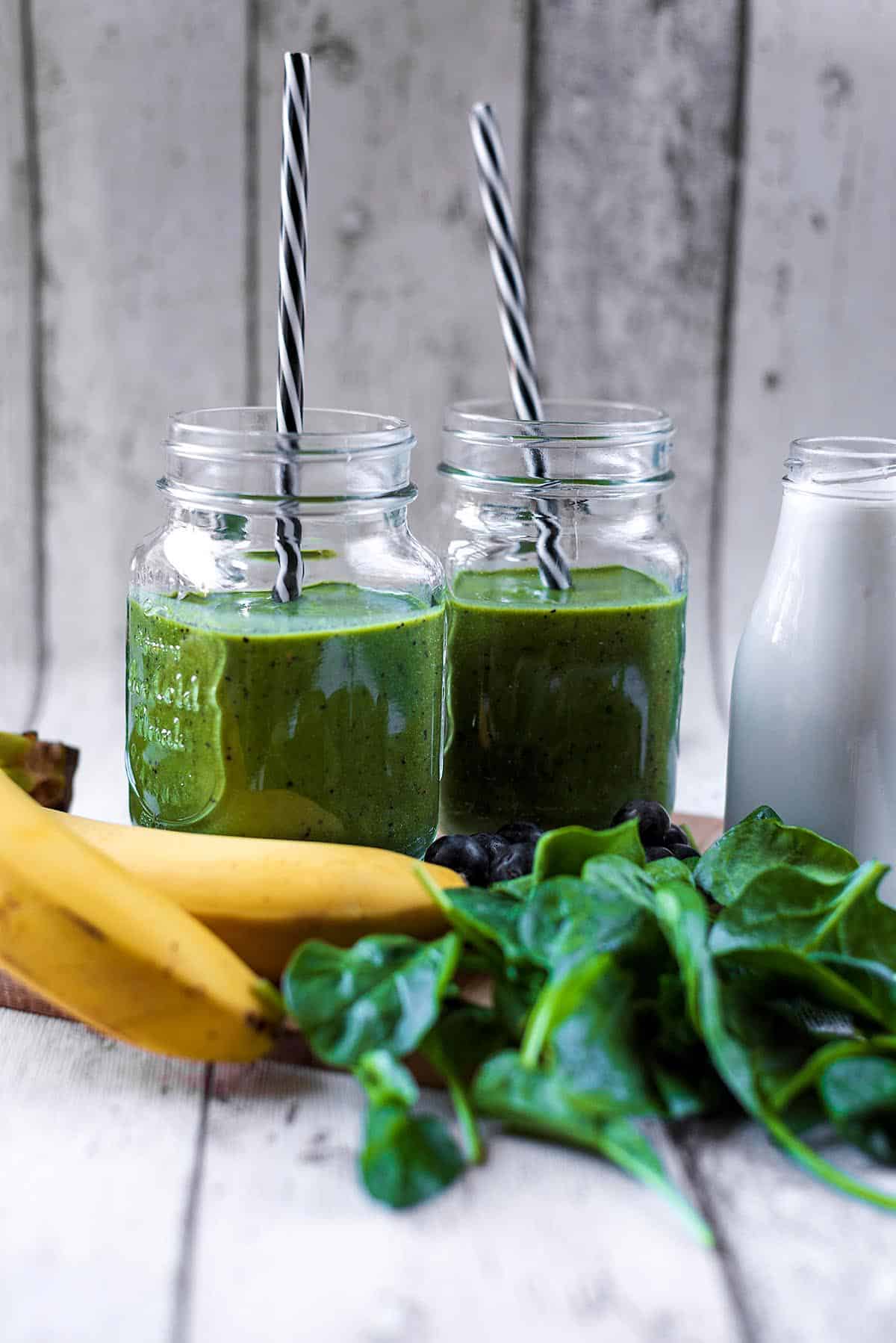 Two jars of green smoothie next to a bottle of milk, some bananas and a pile of spinach.