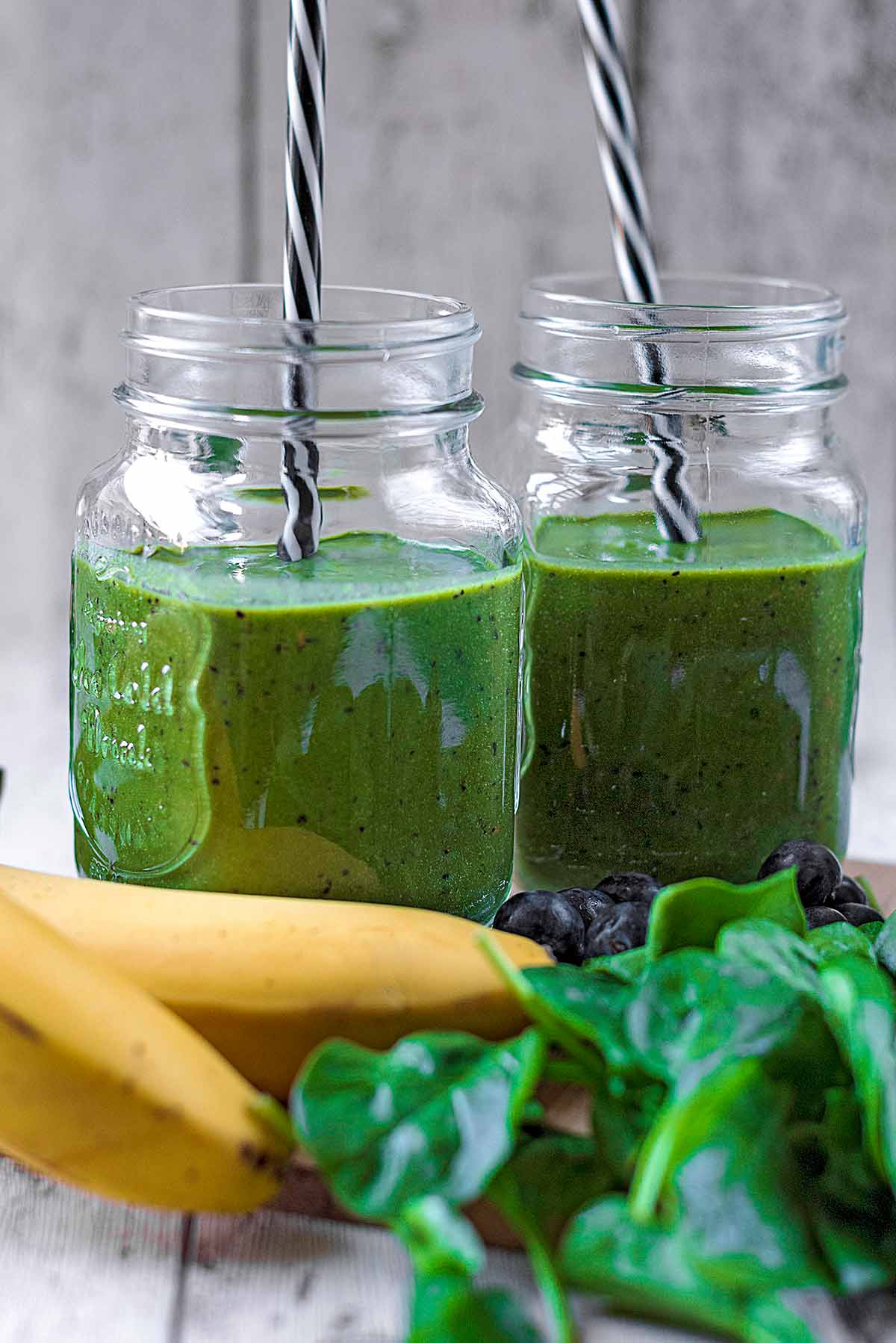 Two jars of green smoothie next to some bananas and a pile of spinach.