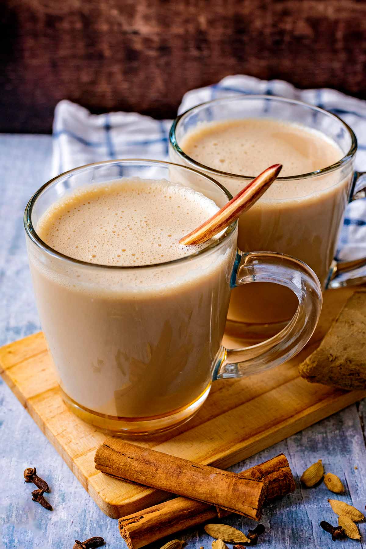 Two classes of chai tea on a serving board next to a cinnamon stick and some cloves.