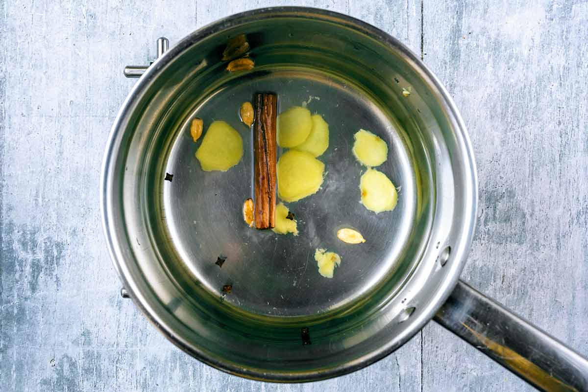 A saucepan containing water, ginger slices, a cinnamon stick, cloves and cardamom pods.
