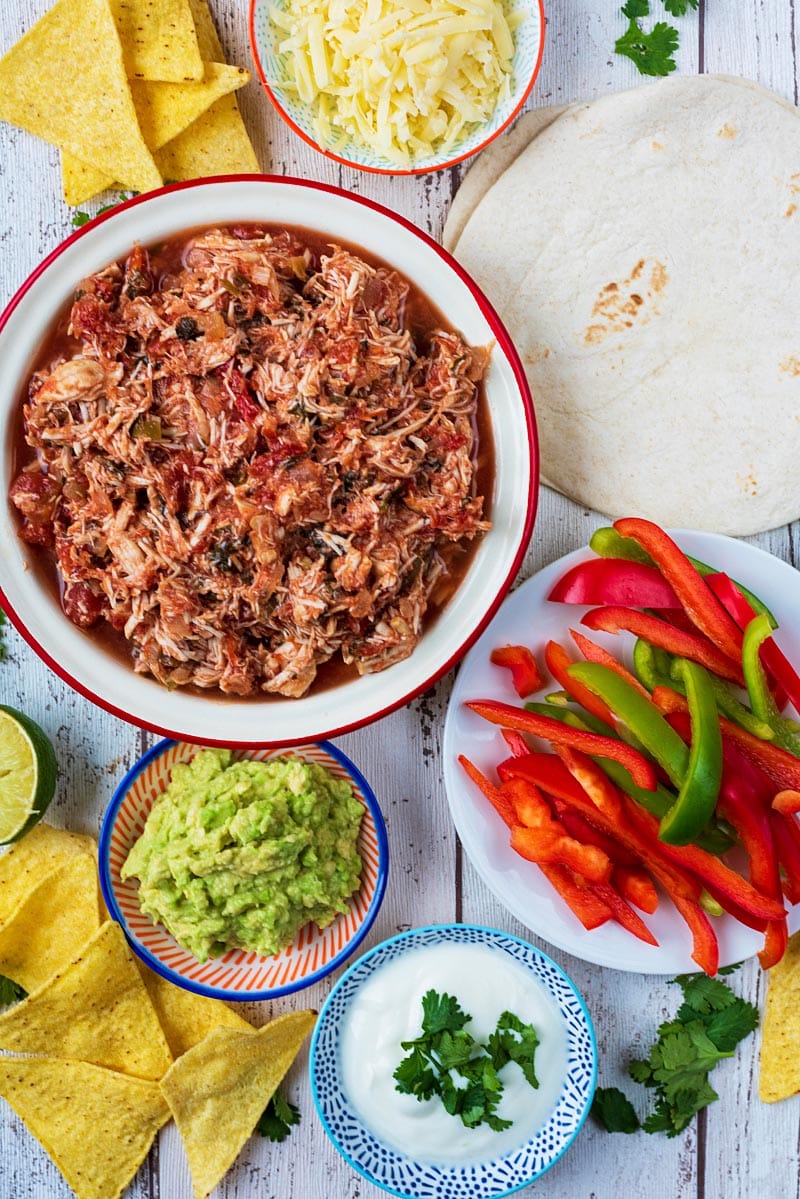 Slow cooker salsa chicken in a dish next to some guacamole, flour tortillas, sliced peppers, sour cream and nachos