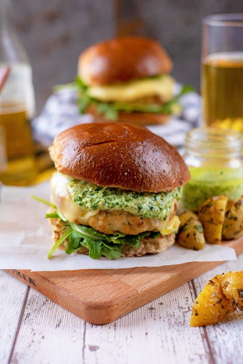 A chicken burger in a bun on a wooden board, beer and another burger in the background