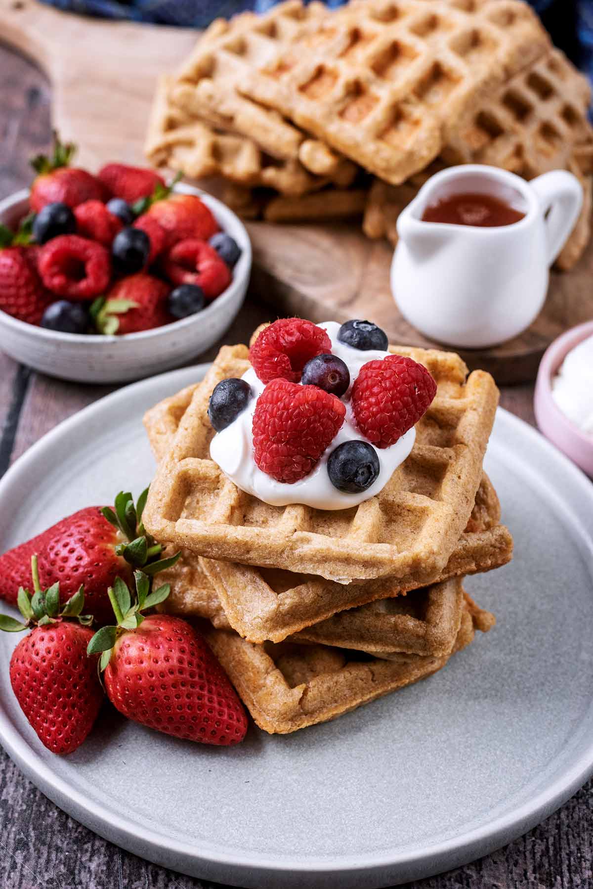 A stack of waffles on a plate with yogurt and berries. More wafflesa re in the background.