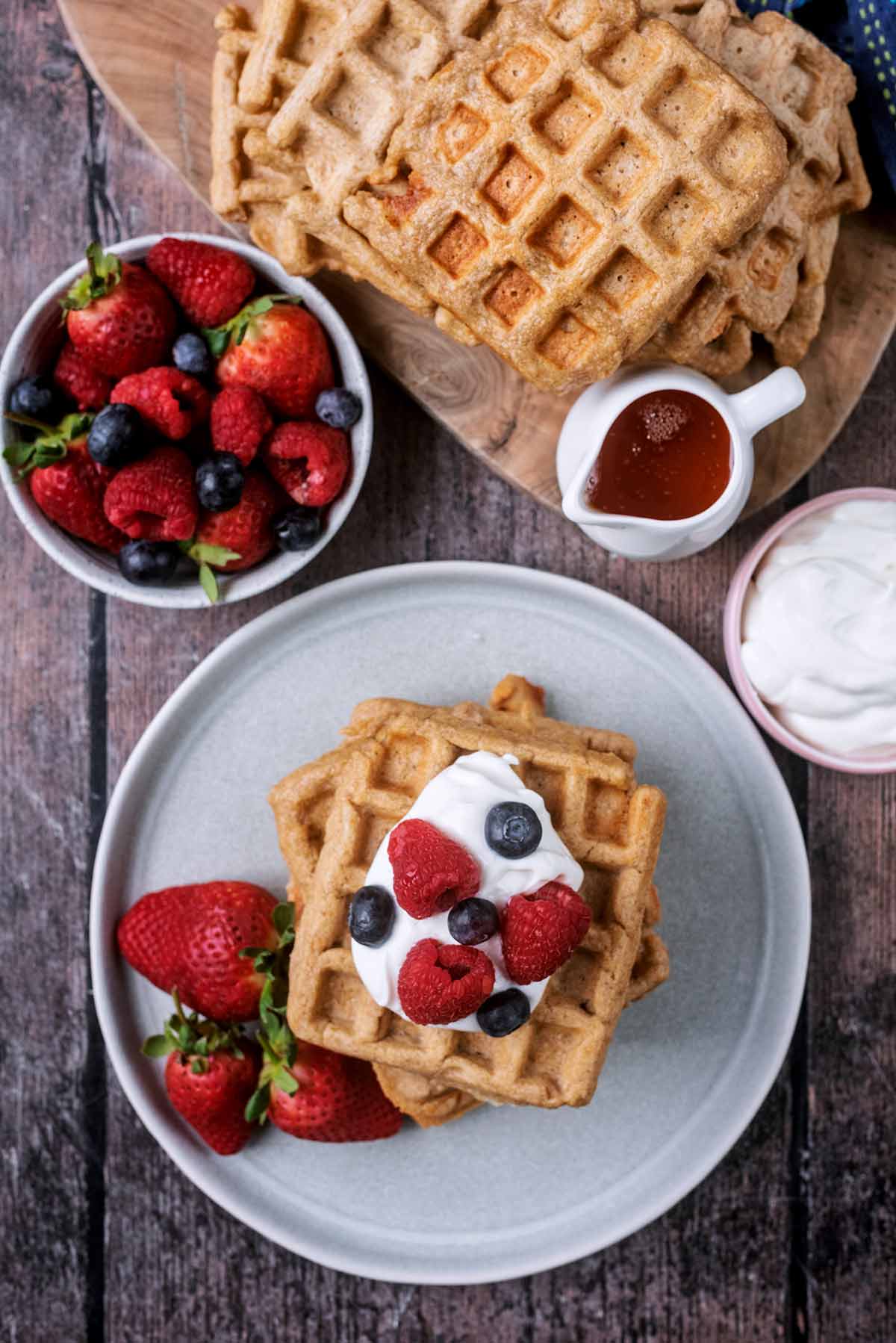 A plate of waffles and berries, next to more waffles, a bowl of berries and a jug of honey.