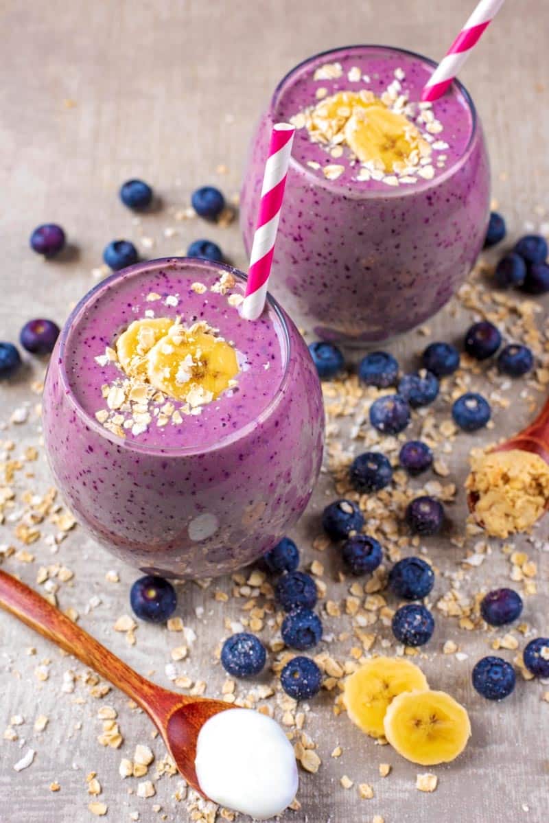 Two glasses of smoothie surrounded by blueberries and oats.