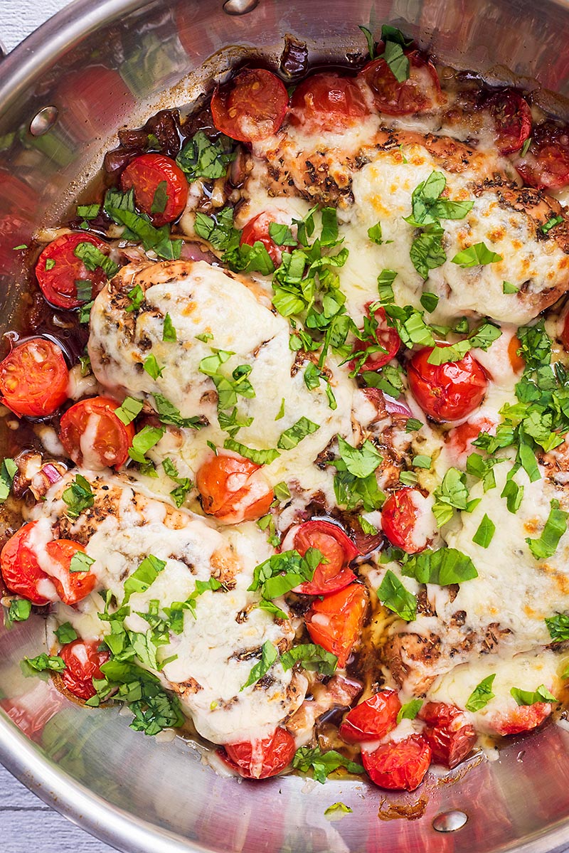 Chicken breasts, covered in melted cheese and chopped herbs, in a large pan with tomatoes.