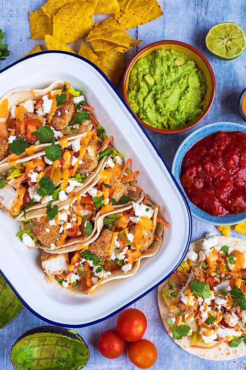 Four Fish Tacos in a rectangular bowl next to bowls of guacamole, salsa and slaw.