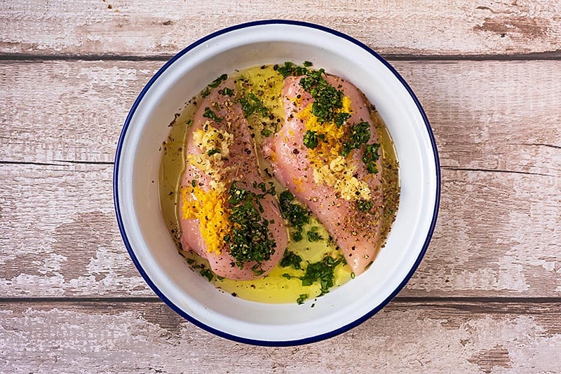 Two raw chicken breasts in a round dish with, lemon juice, zest, oil and chopped basil.